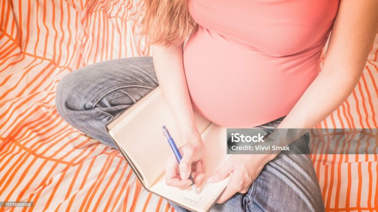 A pregnant redhead woman is preparing for a soon childbirth and handwriting the details of her birth plan in a block note at home.